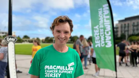Daisy is in a green 'Macmillan Cancer Support Jersey' t-shirt stands outdoors near a large green flag with matching text. A thermometer-shaped sign marked 'Goal' is visible, with other people in the background, indicating an event or gathering