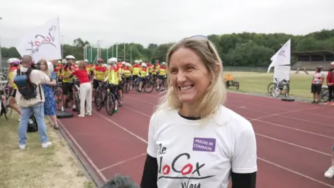 Blonde woman with a white "Jo Cox Way" T-shirt smiles in front of a group of cyclists about to cycle from Cleckheaton to London.