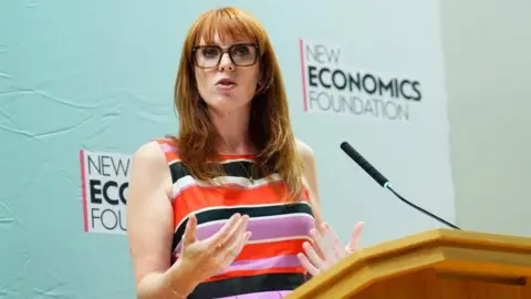 Angela Rayner is stood up and speaking at a wooden lectern. She is wearing a dress with multi-coloured horizontal stripes. She is wearing glasses and has long, ginger-coloured hair. Rayner is stood in front of backdrop which has "New Economics Foundation" written on it.