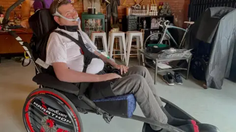 Sam Perkins sat in his race chair. He is wearing a white t-shirt and joggers.