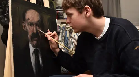 Amanda Dewar Connor Draycott. who has short light brown hair and is wearing a navy jumper, makes finishing touches to his portrait of James Finlayson with a wooden paintbrush.