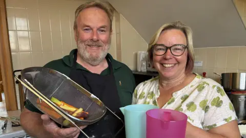A man with short grey hair and a short grey beard is wearing a green polo shirt and holding a tray of sliced vegetables. His is standing next to a woman with short blond hair, glasses and a white and green floral patterned blouse. She is holding turquoise and pink water jugs. Both are smiling
