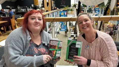 Carys Wiggans, on the left, with red hair, wearing a grey cardigan and a blue top with roses on it. She has a ring on her middle finger and her ring finger. She is Holding a book, titled The Great Theatre Rescue, which is wrapped in green ribbon and has a book fairy sticker on it. Kate Pearson, on the right, wearing a pink jumper, her blonde hair is pulled back. She is wearing a necklace, the pendent is square and is multi-coloured with white, pink and blue. She is wearing a ring on her ring finger. She is holding a green book, with pink ribbon on it and a book fairy sticker, with a bookmark which says 'Free Book' sticking out of the top. 