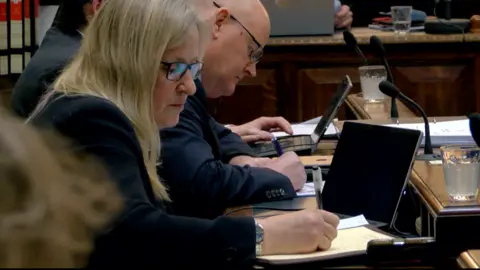 Michelle Haywood, a woman with blonde hair, she sits down and wears glasses, she writes on paper, and there is a laptop in front of her.