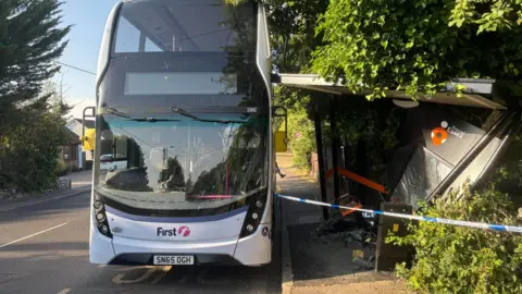 A bus parked a few inches from the kerb, but in contact with the bus shelter which is collapsed. Police tape is blocking off the scene.