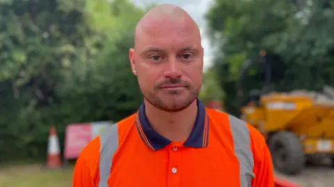 A man wearing a bright orange polo shirt with hi-vis stripes down the shoulders and a blue collar. He has a shaved head and short-cropped stubbly beard. There is construction equipment in the background as well as trees.