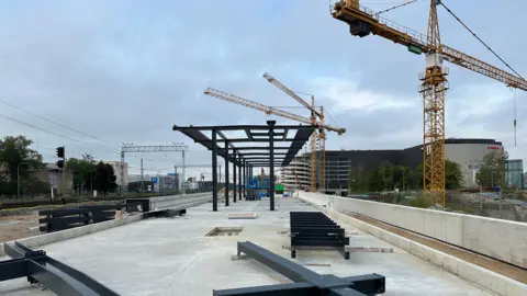 Al frente se extiende una plataforma inacabada, con listones negros apoyados sobre el hormigón y que se elevan en una estructura a medio construir, junto a una grúa.