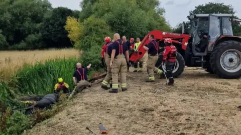 Carly Wilford A tractor with a rope attached to it that is pulling a horse covered in mud from a ditch, surrounded by firefighters in a field. 