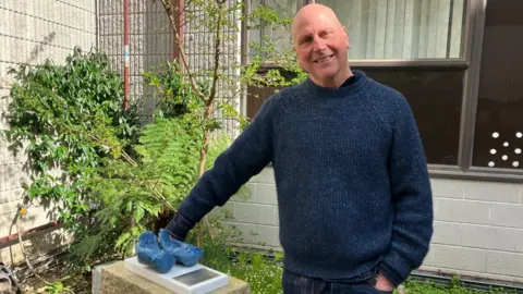 Kurt Jackson is standing in the healing garden on a sunny day with his hand resting on a plinth. On the plinth is The Nurse's Hands sculpture. It is a pair of cupped hands in a blue material. He is smiling and is wearing a blue woolly jumper and jeans. His other hand is in his pocket.