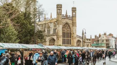 Beverley Minster with market stalls with green and white roofs in front. Crowds of people fill the road where the stalls are.