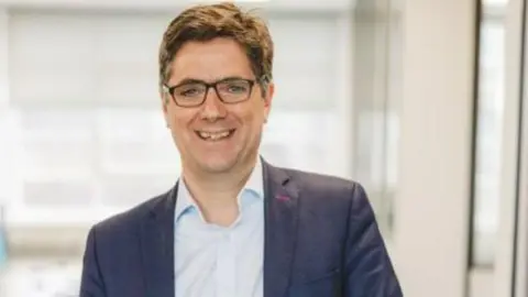 Aidan Dunn is smiling at the camera. He has short brown hair and wears thick plastic framed glasses. He has a pale blue collared shirt on and a navy blue blazer.