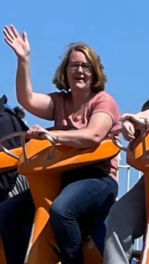 Family Handout Tracy Godfrey waves, she is wearing a pink t-shirt and jeans. She's sat on what looks like an orange roller coaster seat. 