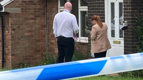 Police on a doorstep of a property at the scene of a crime, behind blue and white tape