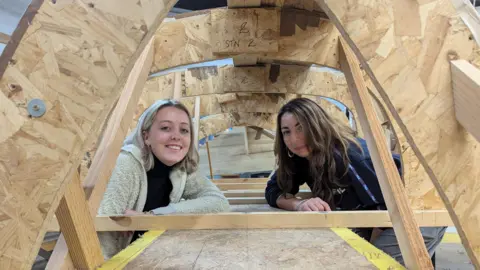 Sophie Harding (left), who has shoulder length blonde hair and a white fleece, and Hannah Lovett (right), who has shoulder length brown hair, sat beneath the shell of a boat currently under construction