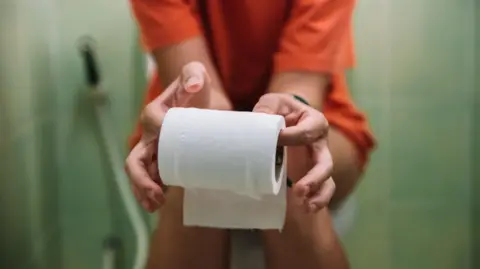 A blurred woman sits on the toilet in a green room holding a white toilet roll. She is wearing an orange shirt.