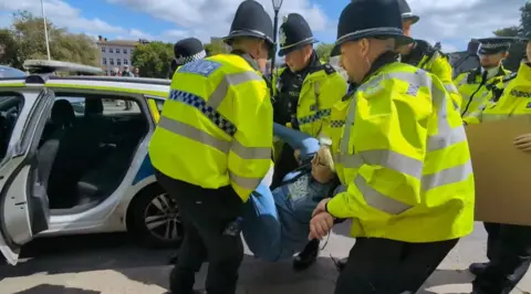 Claire Bullion Several police officers carry a woman in blue jeans and sweatshirt towards the open door of a police car.