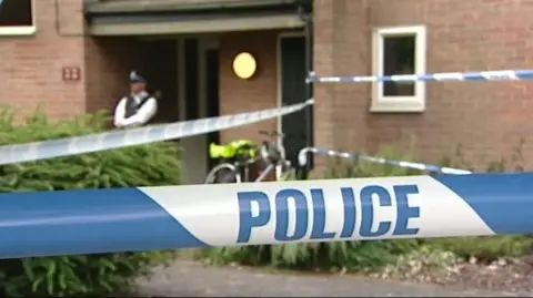 A still of an archive video from the time of the murders. It shows a police officer stood outside a property with police going from one side to the other.