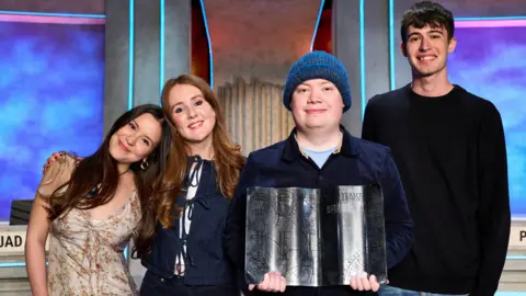 A group of four people stand side by side - left to right, a woman with long dark hair and a pale flowery dress who has her head on the shoulder of a woman with long red hair and wearing a blue top, her arm around the other woman, a person in a blue beanie and blue top holding a silver award in the shape of an open book, and a man with short dark hair and a dark jumper. 