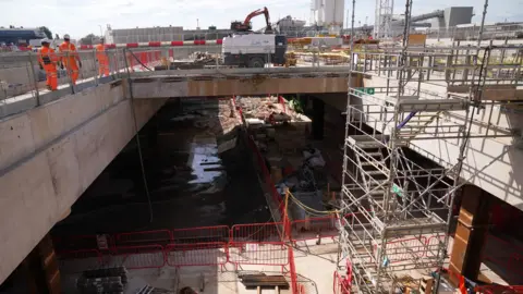 PA Media A general view of the HS2 site at Old Oak Common, in west London, showing a concrete underpass with metal scaffold tower on the right from the ground up, and on the bridge section, there are three workers in orange hi-vis suits. A yellow crane can be seen in the background and the London skyline of buildings. Picture date: Thursday August 10, 2023.