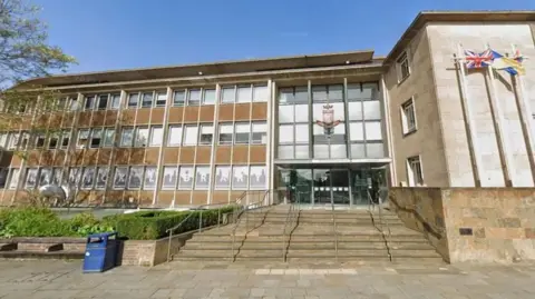 A large light brick building with a glass entryway, there is a wide set of stairs leading up to the building