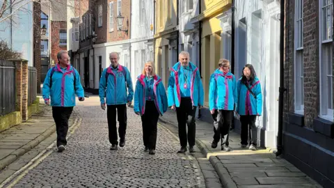 Four men and two women, all wearing bright blue jackets and black trousers, walk in a row down a cobbled, curved street with Georgian terraced houses, painted in various colours, to one side.