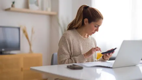 Woman sits at a desk in a home with a phone and a laptop in front of her and a calculator in her hand.