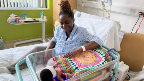 Jessica Bayley/BBC A woman wearing a light blue shirt is sat on a hospital bed smiling down at her son beside her who is in a clear baby cot and has a colourful crochet blanket on top of him.