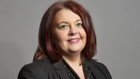 UK Parliament Headshot of Liverpool Wavertree MP Paula Barker. She has shoulder length dark, auburn hair and is wearing a black jacket and a black top with small yellow spots.
