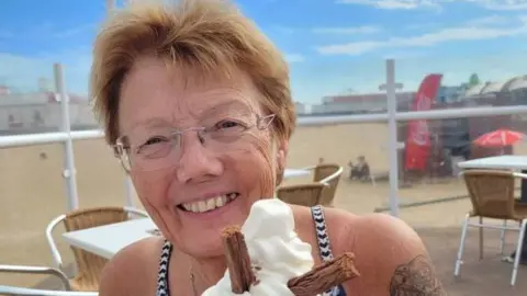 Penny Bulpitt: a woman with short brown hair wearing glasses and smiling. She is holding an ice cream cone. Behind her are tables, chairs and a beach.