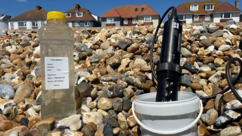 A plastic water bottle is full of seawater on a beach with a white label on it
