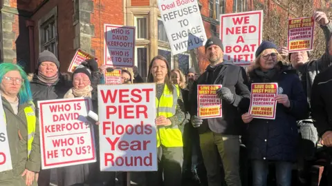 Men and women stand with placards that read "Who is David Kerfoot?", "we need a boat lift" and "West Pier floods year round".