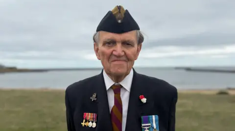 BBC/Jim Scott Rodney Buckton, who is now an Army veteran, is wearing a blue beret with a mustard yellow addition. He is also wearing a blue blazer with a variety of medals from his time in the Army being displayed on both his left and right hand side. In the background is the mouth of the River Tyne, along with both north and south piers, which looks gloomy because of the overcast skies.