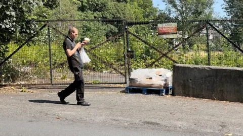 Aisha Iqbal/BBC A man walking past what looks like a derelict site, overgrown with shrubbery and fenced off.