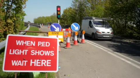 Jamie Niblock/BBC A van passes the traffic lights on Woodend Bridge in Witham. One of the lanes is blocked off by cones, signage and a traffic light.
