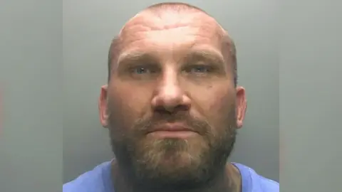 Mugshot of Derek Heggie, who is bald with a broad build and wearing a blue top. He has a short beard and moustache flecked with lighter coloured hairs.