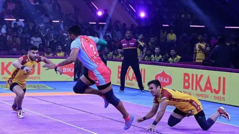 Kabaddi match in India in 2024. One player is jumping while two opposing players reach towards him. There is a referee in the background and audience members are sitting and standing. The match is illuminated by bright lights.