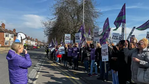 Unison People in purple jackets standing outside the hospital. Lots of people holding purple flags and holding signs: "Pay Fair". 