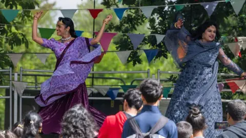 Ipswich Borough Council Two women in purple saree dresses dance on stage with their arms in the arm. A crowd watches them. Bunting has been placed across the back of the stage. 