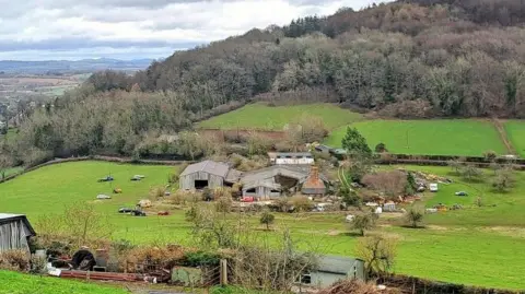 LDRS A view of the farm from the immediate roadside. The farm sits in the middle of fields and is surrounded by trees.