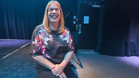 Zoe Wickham sitting on the edge of a stage in a performance hall or theatre. She has her hand on one knee and is smiling at the camera. She has long blonde hair.