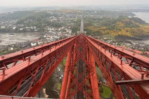 Odran Doherty Looking down the red steel frame of the rail bridge. In the background is hills and houses in Fife.