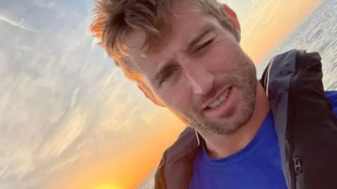 Tom Waddington Tom Waddington taking a selfie with his left eye shut, wearing a blue T-shirt and black life jacket with a sunset and the ocean behind him