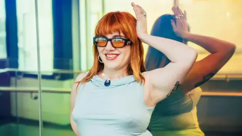 CMAT smiles for the camera while in a studio, standing in front of a mirrored wall. She has red hair and is wearing a pale aquamarine top.