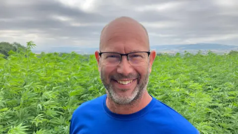 Jake Freestone pictured standing in front of a field of hemp. He has a bald head and grey stubble. He is wearing think glasses and a blue t-shirt.