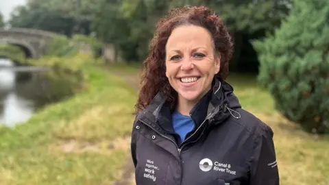 Head-and-shoulders outdoor image of Angela Parkinson-Green standing beside a canal wearing a dark waterproof jacket zipped up to the neck over a blue top. The canal runs along the left side of the image.