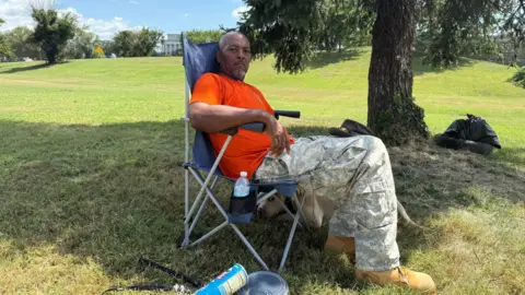 A man in a red t-shirt and camouflage pants sits in a blue camping chair in the shade of a tree on a patch of grass. A bottle of water is in the cup holder. His dog can be seen sleeping by his side, underneath the chair.