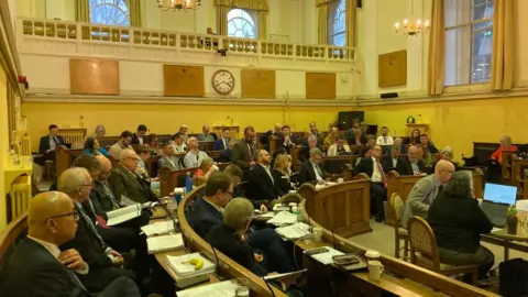 A view of the council chamber from the ground floor. There's a mix of men and women with papers on their desks.