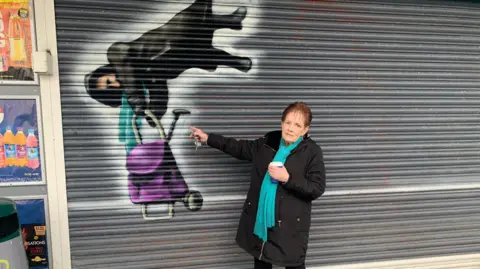 Anne Hughes, with red hair, a black coat and a blue scarf, standing next to a mural of her viral moment on the closed metal shop shutters. 