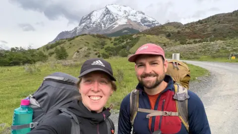Sophie Collier Sophie Collier and Jack Gunter wearing hiking gear including large rucksacks take a selfie while standing in front of a mountain.