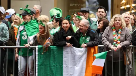 Getty Images A number of people with green hats and head pieces on, and holding Irish flags as they stand behind a barrier watching the St Patrick's Day parade in New York.